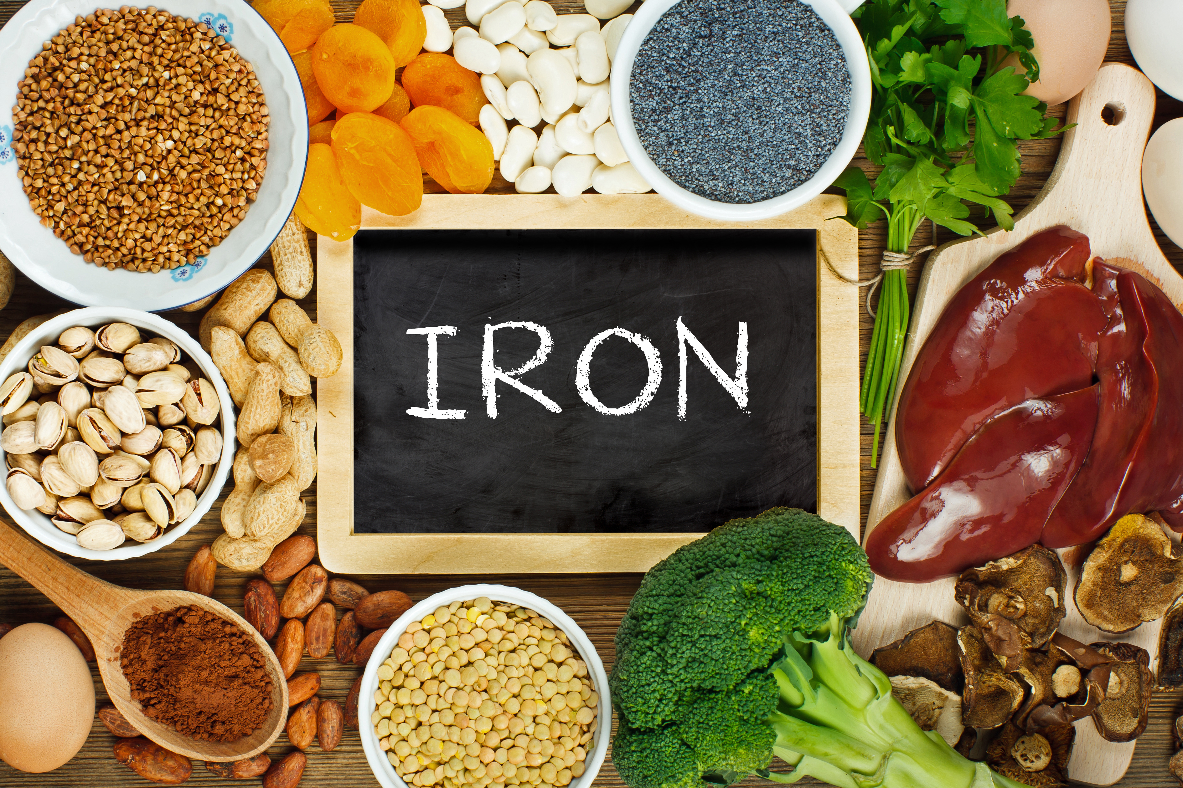 Collection of iron rich foods such as liver, buckwheat, eggs, parsley leaves, dried apricots, cocoa, lentil, bean, blue poppy seed, broccoli, dried mushrooms, peanuts and pistachios on wooden table.