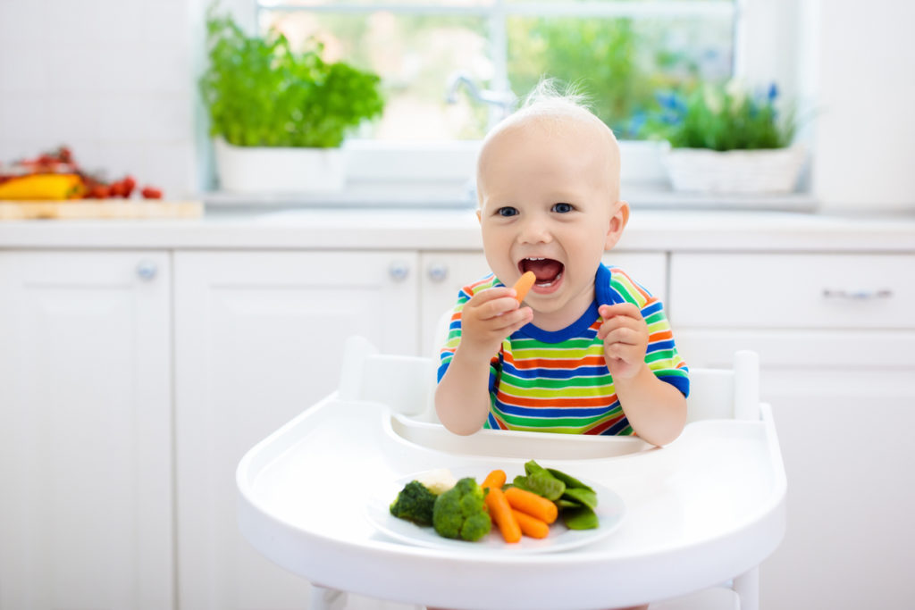 Cute baby eating a solid food, organic broccoli, cauliflower, carrot and green peas. in white kitchen.