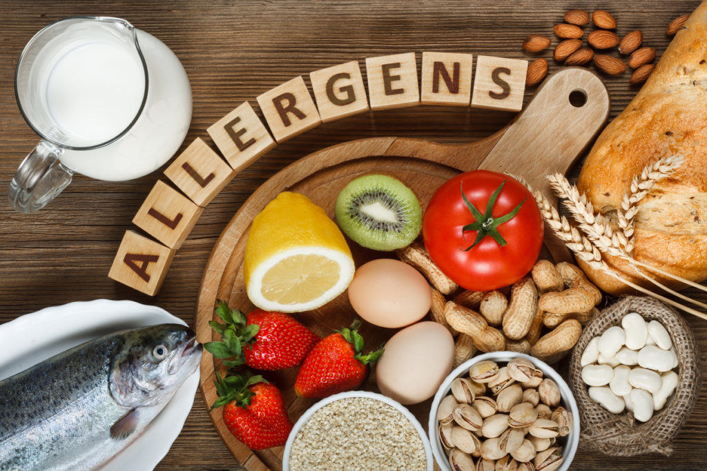 Allergen foods such as almonds, milk, pistachios, tomato, lemon, kiwi, trout, strawberry, bread, sesame seeds, eggs, peanuts and bean on a wooden plate.