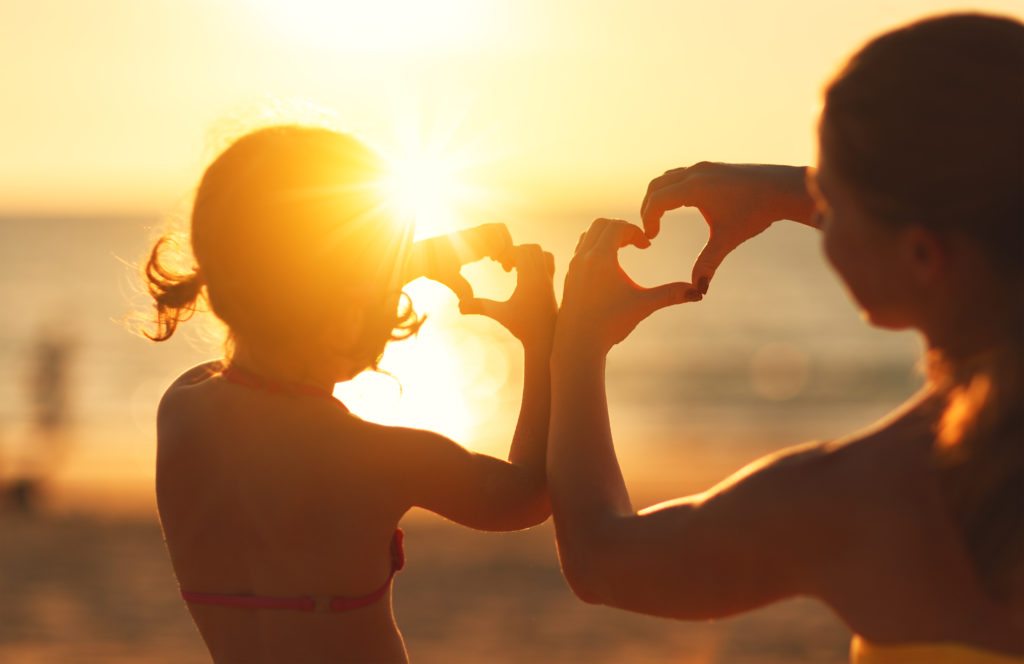Mother and child daughter show heart from hands at sunset on beach.